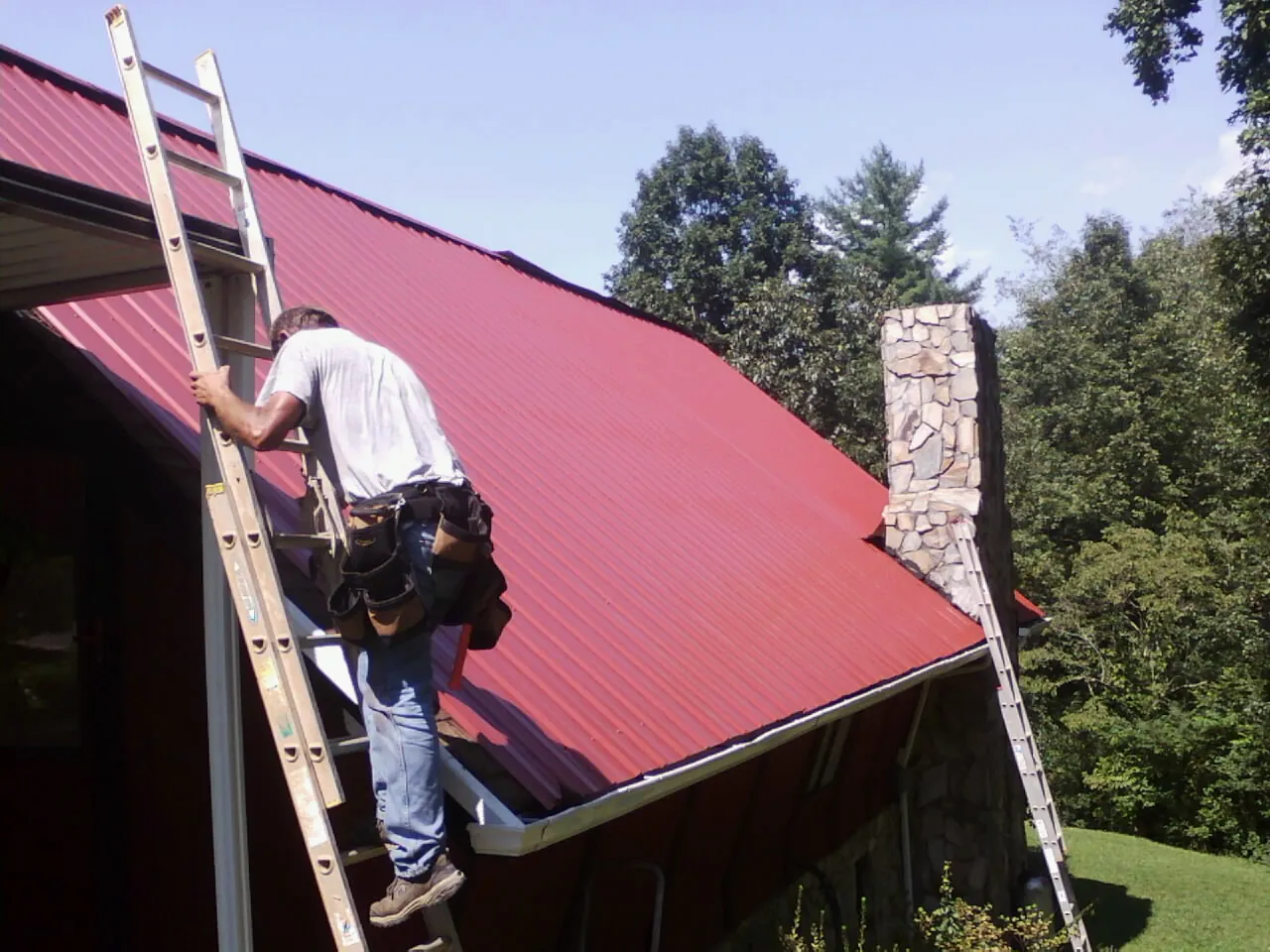 Roofer on a ladder beside a red metal roof and stone chimney — Soffit & Fascia Repair by Roofing Brothers in Martinsburg